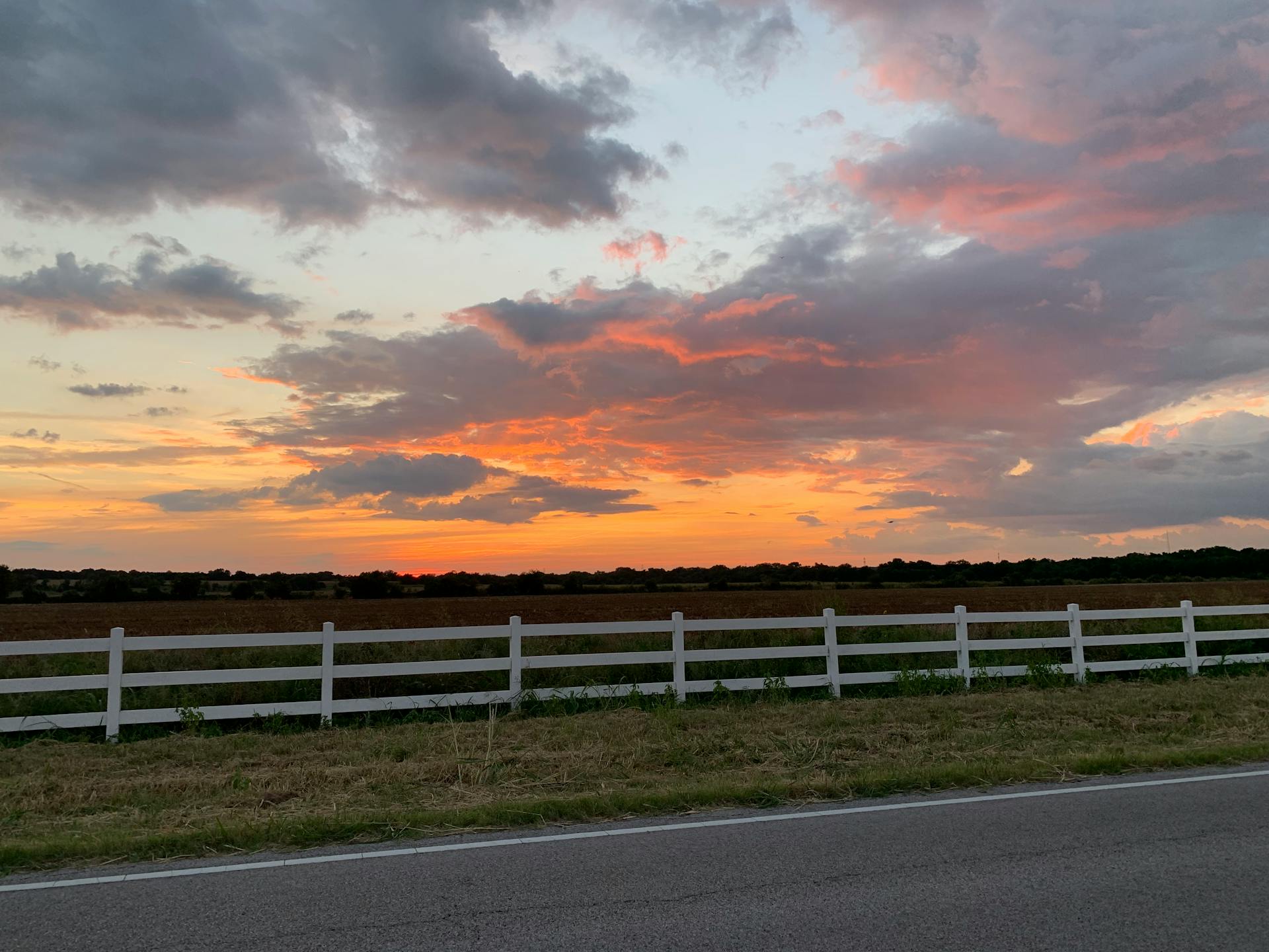 A colorful sunset over a rural landscape with a white fence and road.